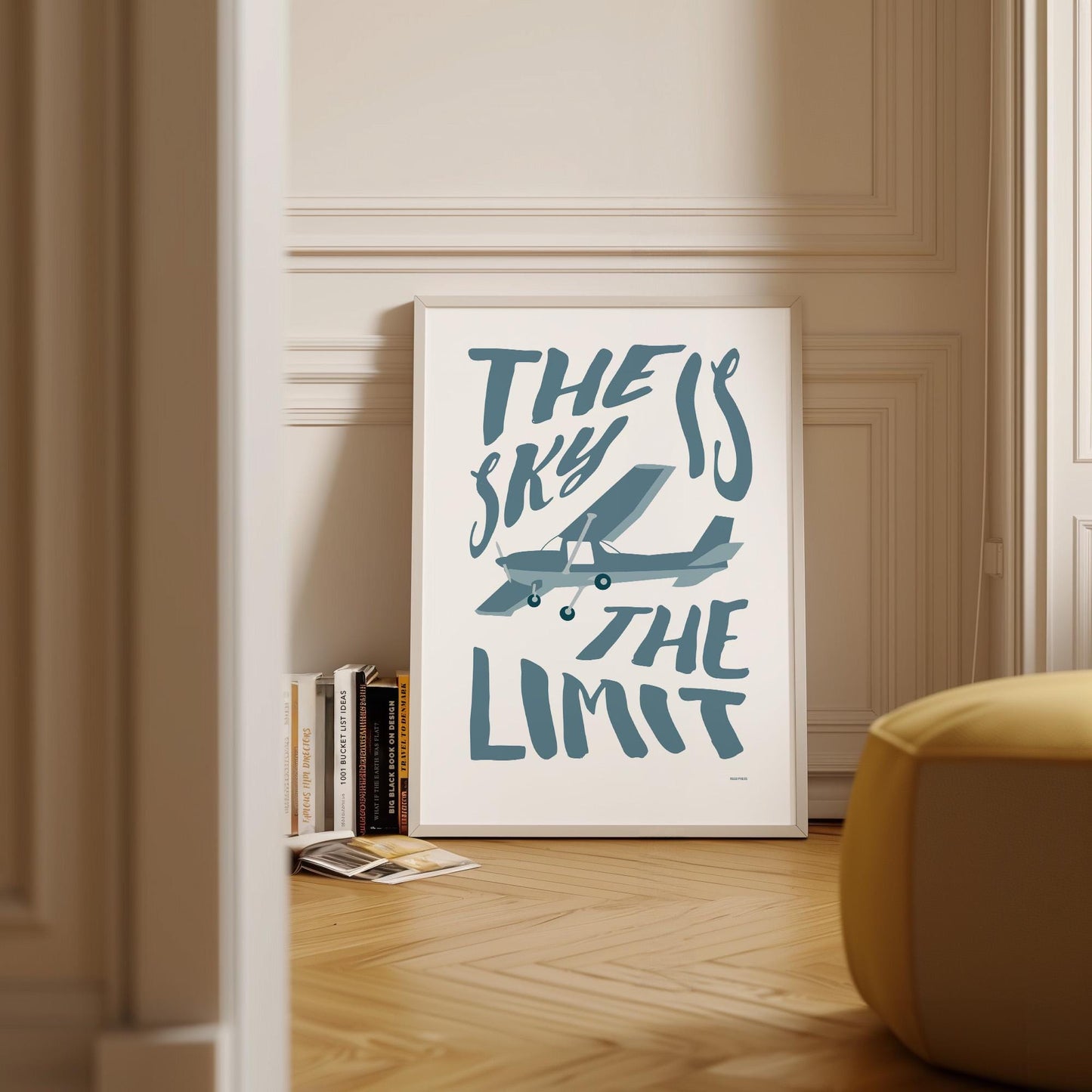 Framed poster on wood flooring leaning against a wall with Scandinavian decor and books around it. It shows an illustration of a light aircraft with the words THE SKY IS THE LIMIT composed around it in blue