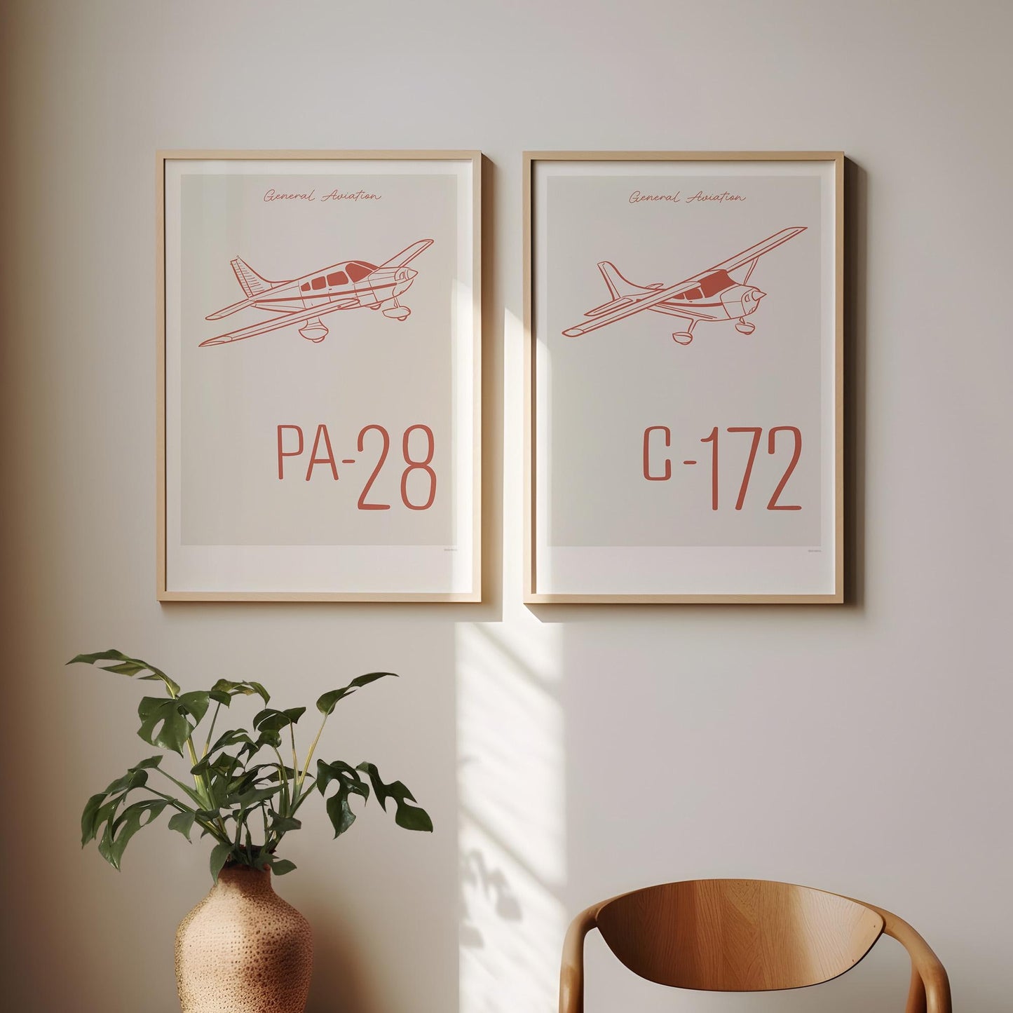 Two framed posters hanging on a white wall above a chair and a plant. Each shows a light aircraft illustration with the words General Aviation and the aircraft name in red