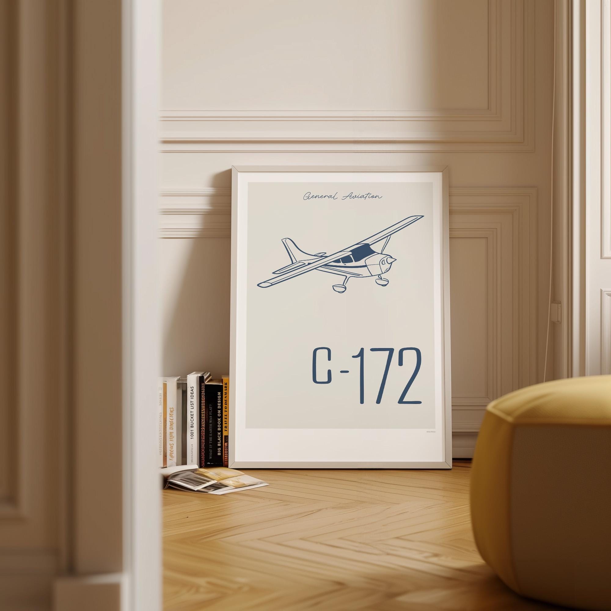 Framed poster on wood flooring leaning against a wall with Scandinavian decor and books around it. It shows a light aircraft illustration with the words General Aviation and C-172 in navy blue